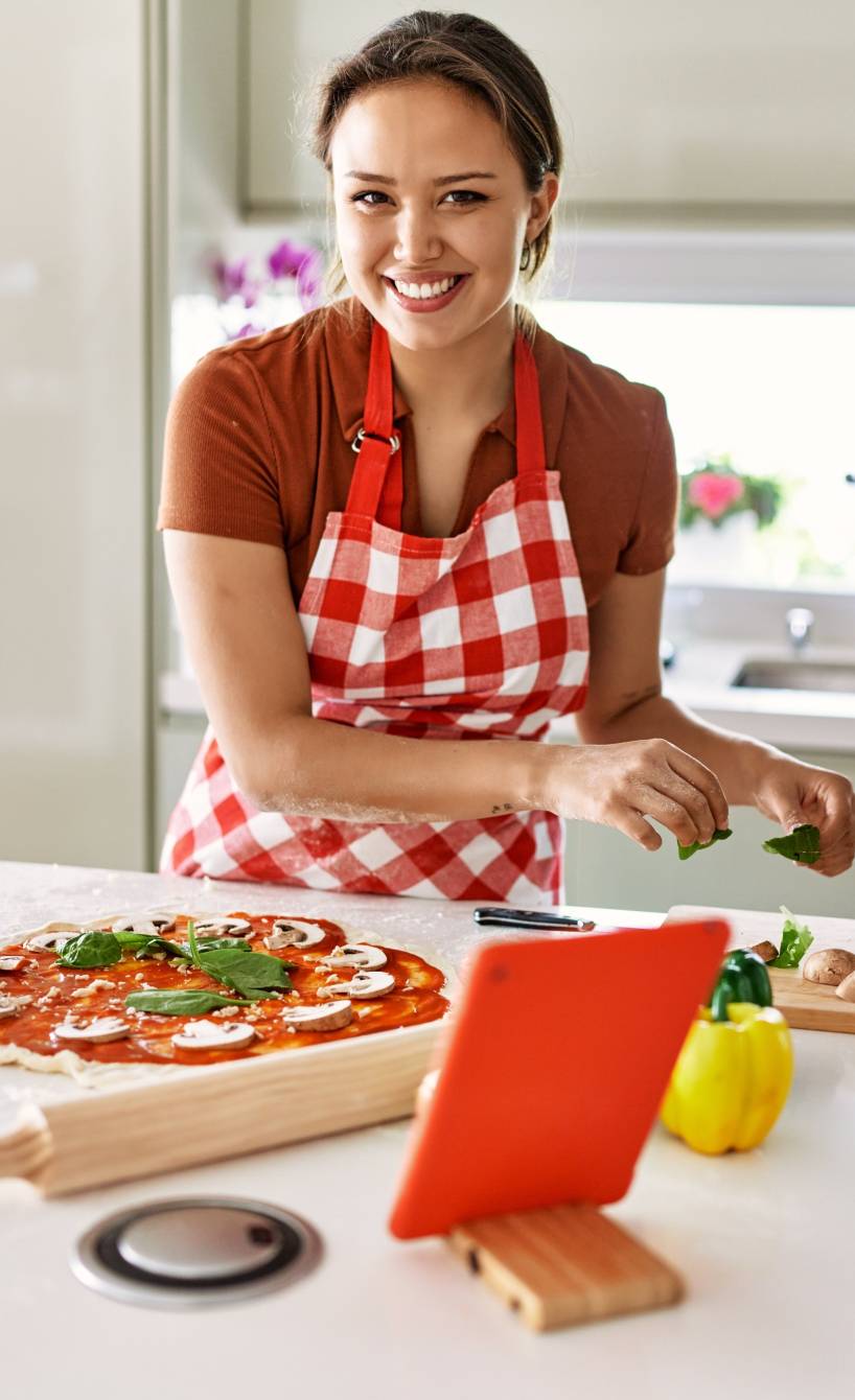 Eine Frau steht hinter einer Kücheninsel, auf der sich Zutaten für Pizza sowie ein Tablet zum Lesen des Rezeptes befinden
