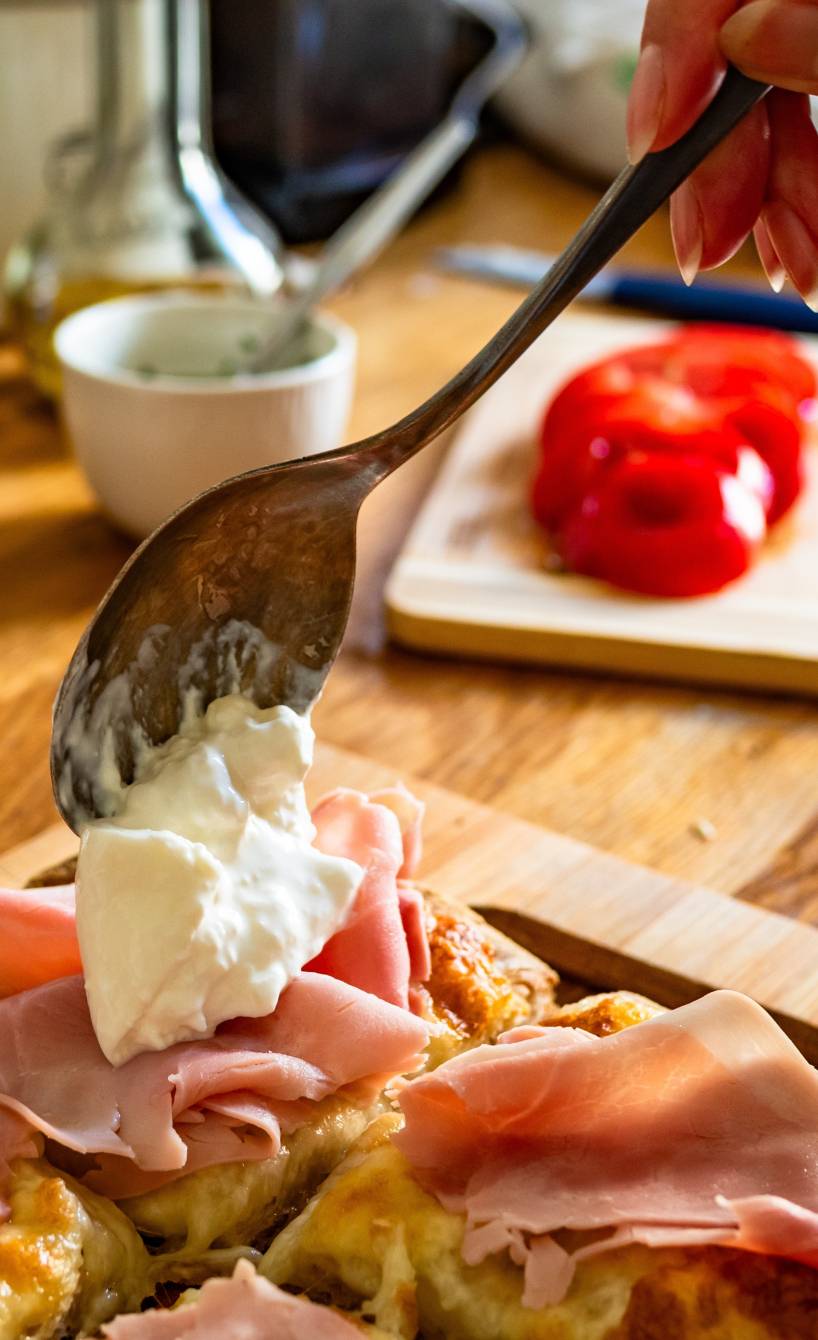 Jemand toppt eine belegte Pinsa mit Burrata, drumherum Zutaten wie Tomaten und Schinken