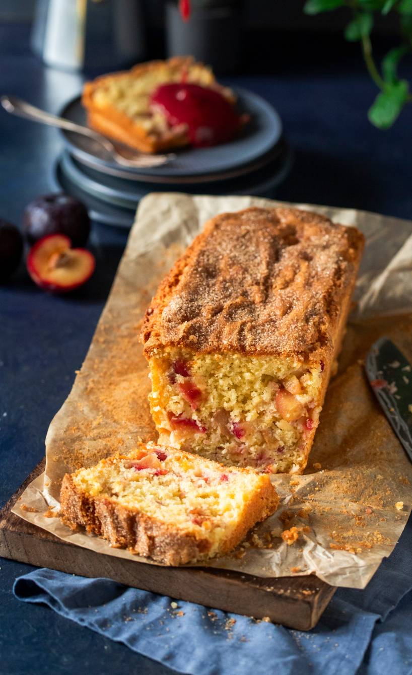 Pflaumen-Olivenöl-Kastenkuchen angeschnitten auf einem Holzbrett, dahinter wird gerade ein Stück mit Pflaumensoße übergesossen