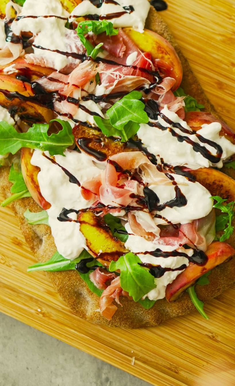 Gebackene Pinsa belegt mit gegrilltem Pfirsich, Burrata und Parmaschinken auf einem Holzbrett
