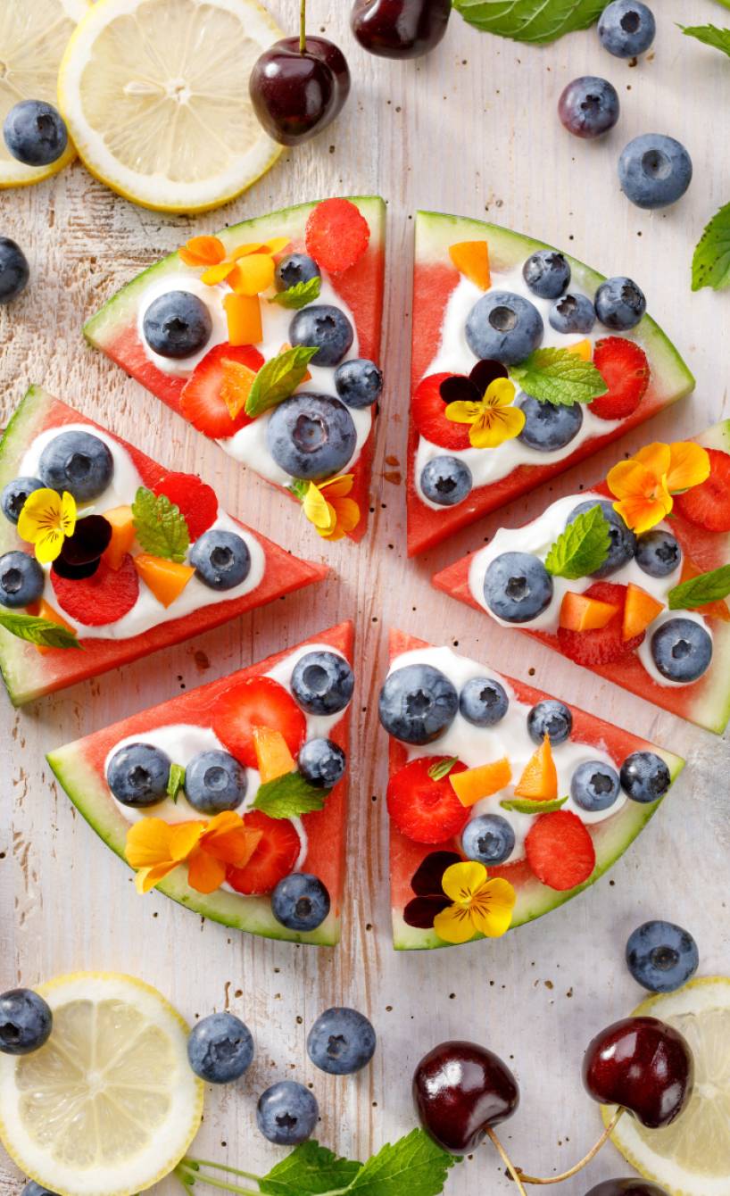 Wassermelonen-Pizza aus der Vogelperspektive, bestrichen mit weißer Ricotta-Creme und bunt belegt mit Blaubeeren, Erdbeeren, Aprikosen, frischer Minze und essbaren Blüten auf hellem Holzhintergrund