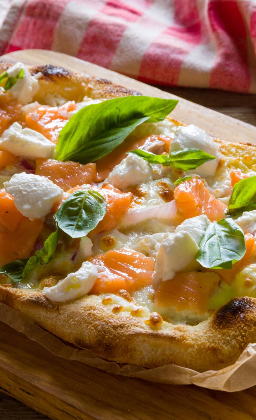 Ganze gebackene Pinsa Bianca mit Lachs und Ricotta getoppt von frischen Basilikumblättern auf einem Holzbrett