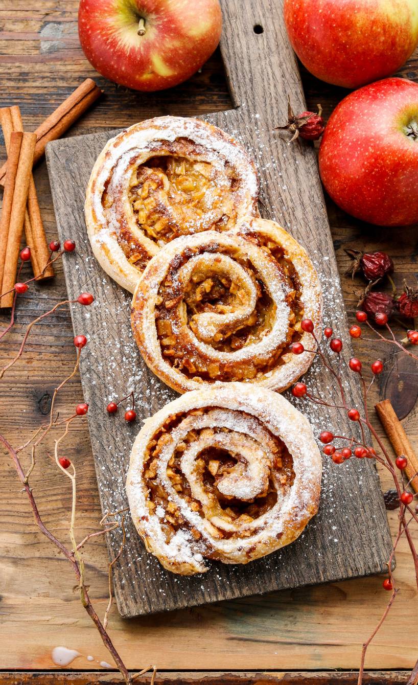 Drei selbstgebackene Apfel-Zimt-Schnecken angerichtet auf einem Holzbrett, daneben frische Äpfel