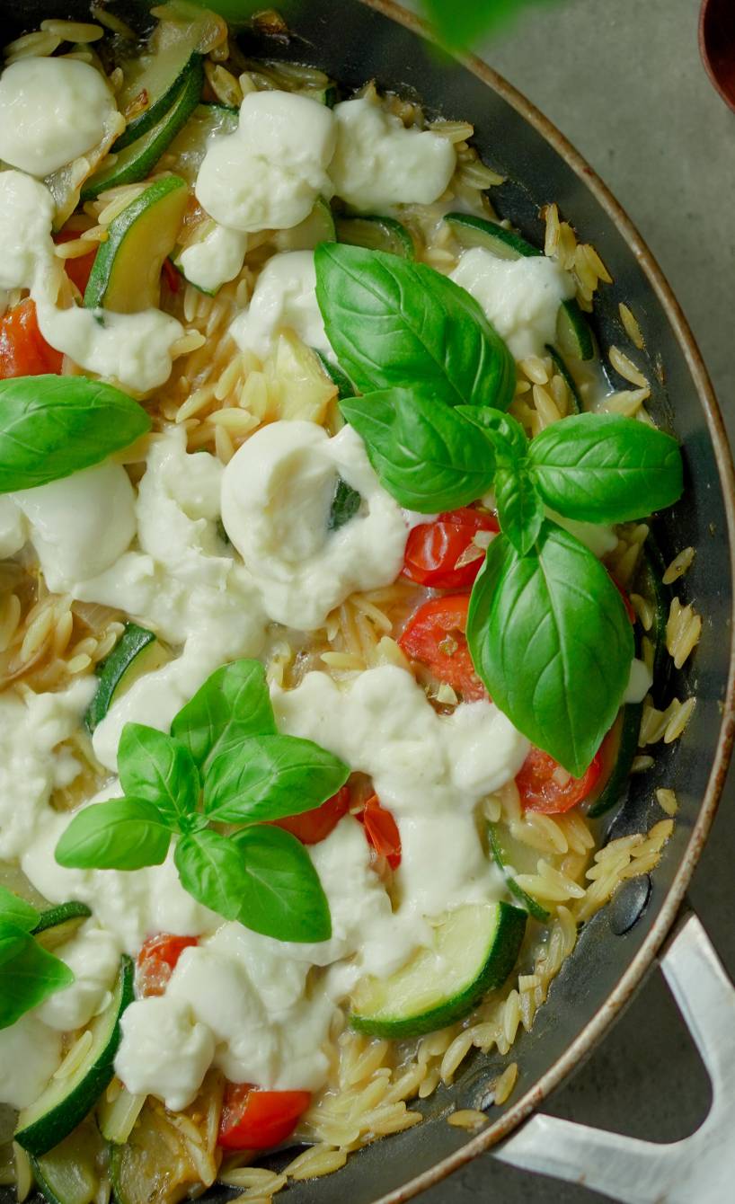 Pfanne mit One-Pot Risoni mit Zucchin und Tomaten getoppt von geschmolzenem Mozzarella und frischen Basilikumblättern