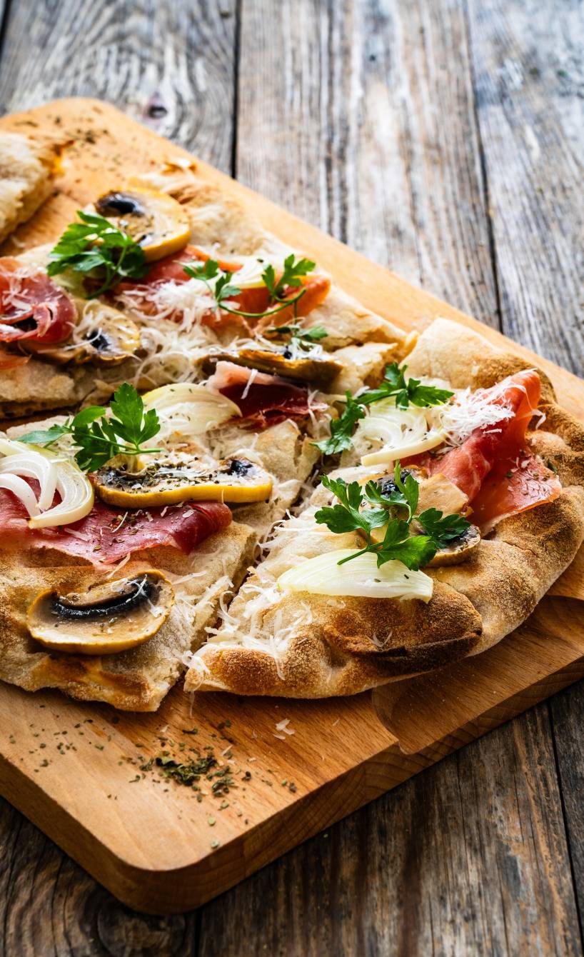 Pinsa Bianca belegt mit Champignons, Mozzarella und Parmaschinken in Streifen geschnitten auf einem Holzbrett