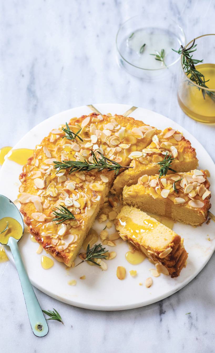 Runder, angeschnittener Polentakuchen mit Mandeln und Ricotta übergossen mit Honig-Rosmarin-Sirup auf einer Kuchenplatte