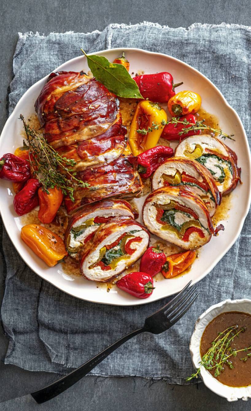 Mediterraner Putenrollbraten im knusprigen Speckmantel angeschnitten auf einer Servierplatte mit bunten Mini-Paprika, daneben Bratensauce