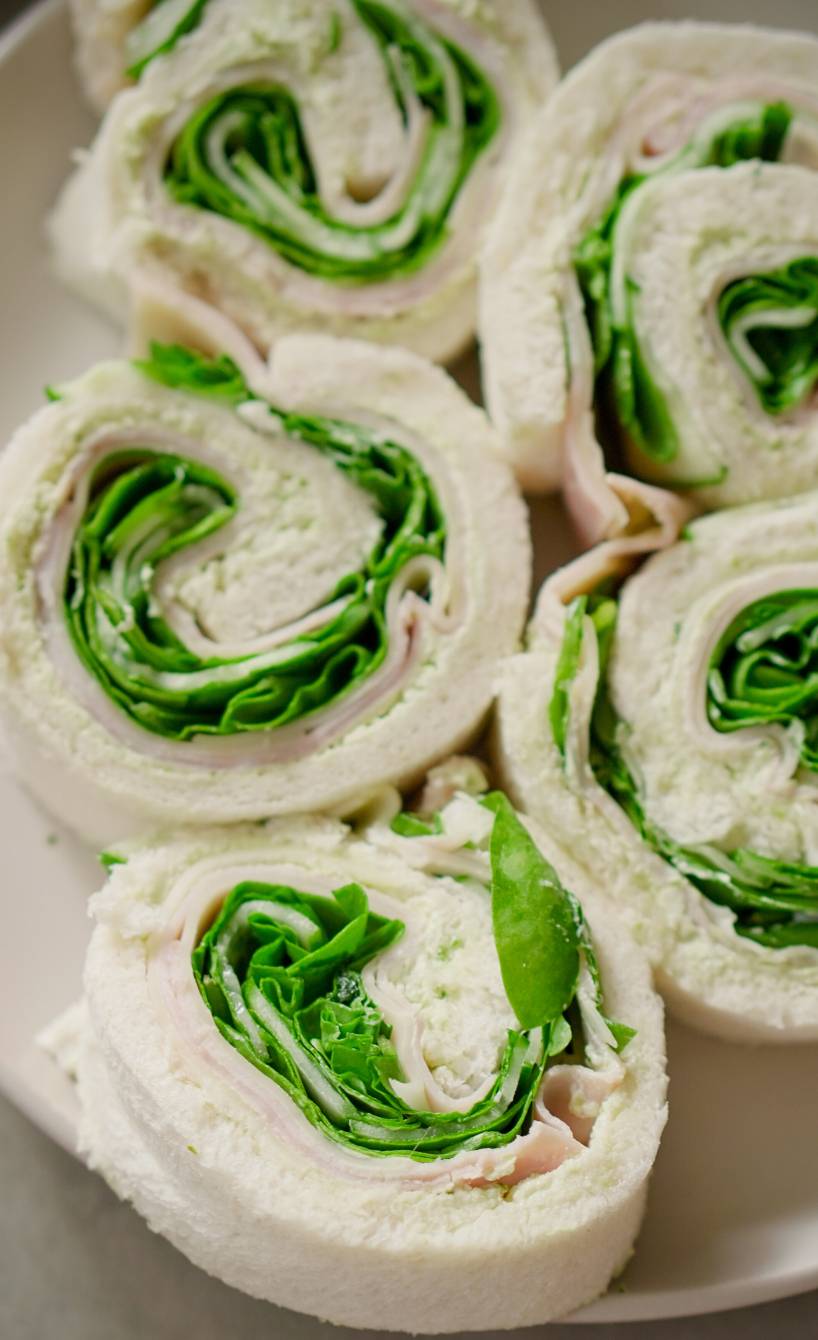 Mehrere selbstgemachte Tramezzini-Röllchen mit Putenbrust. Salat und Frischkäse angerichtet auf einer Fingerfood-Platta und in einer Brotdose