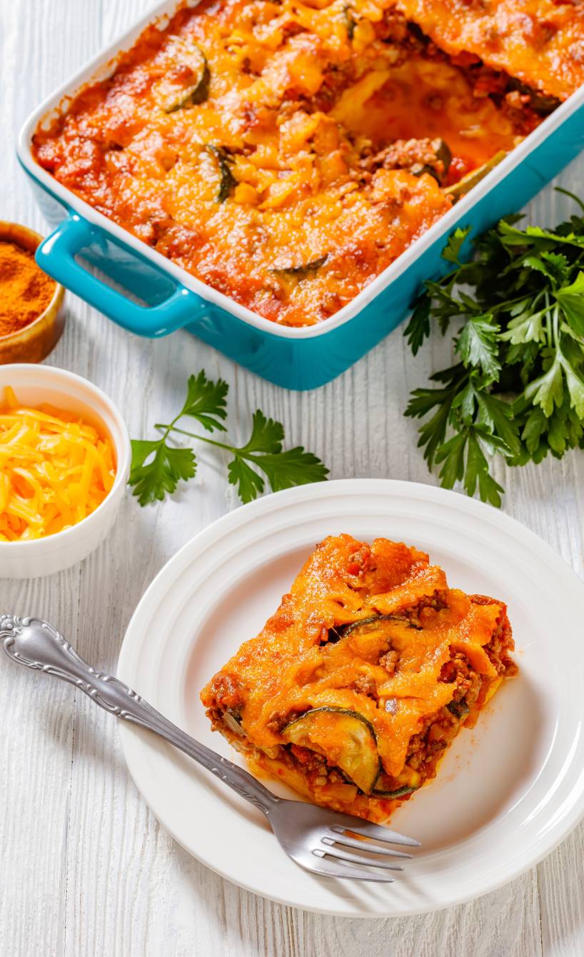 Ein Stück Polenta-Auflauf mit Hackfleisch und Zucchini auf einem Teller, dahinter die Auflaufform auf einem Tisch, daneben frische Petersilie