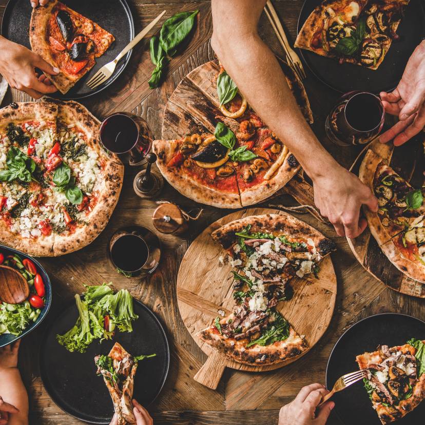 Ein Tisch von oben mit vier verschieden belegten Pizzen, die von mehreren Personen aufgeteilt werden
