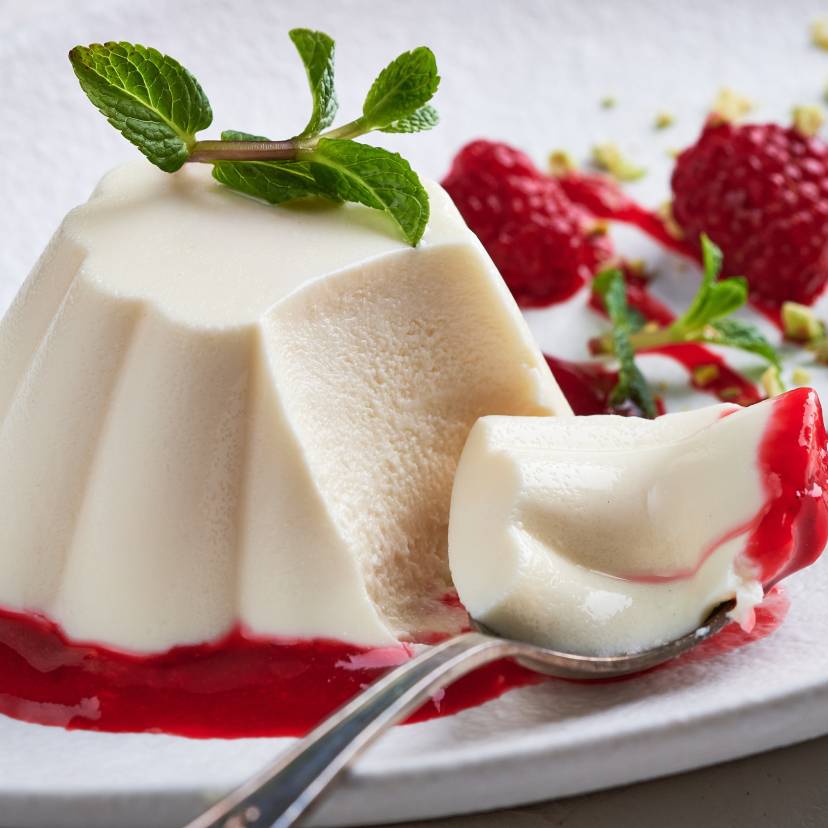 Panna Cotta auf einem Spiegel aus Himbeersoße, daneben frische Himbeeren und Minzblätter